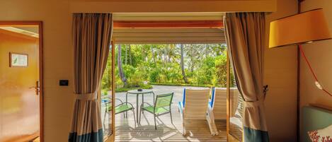 A room with a wooden ceiling, a bed with a blue mattress and a decorative pillow, a table with chairs, and a view of greenery through the curtains.