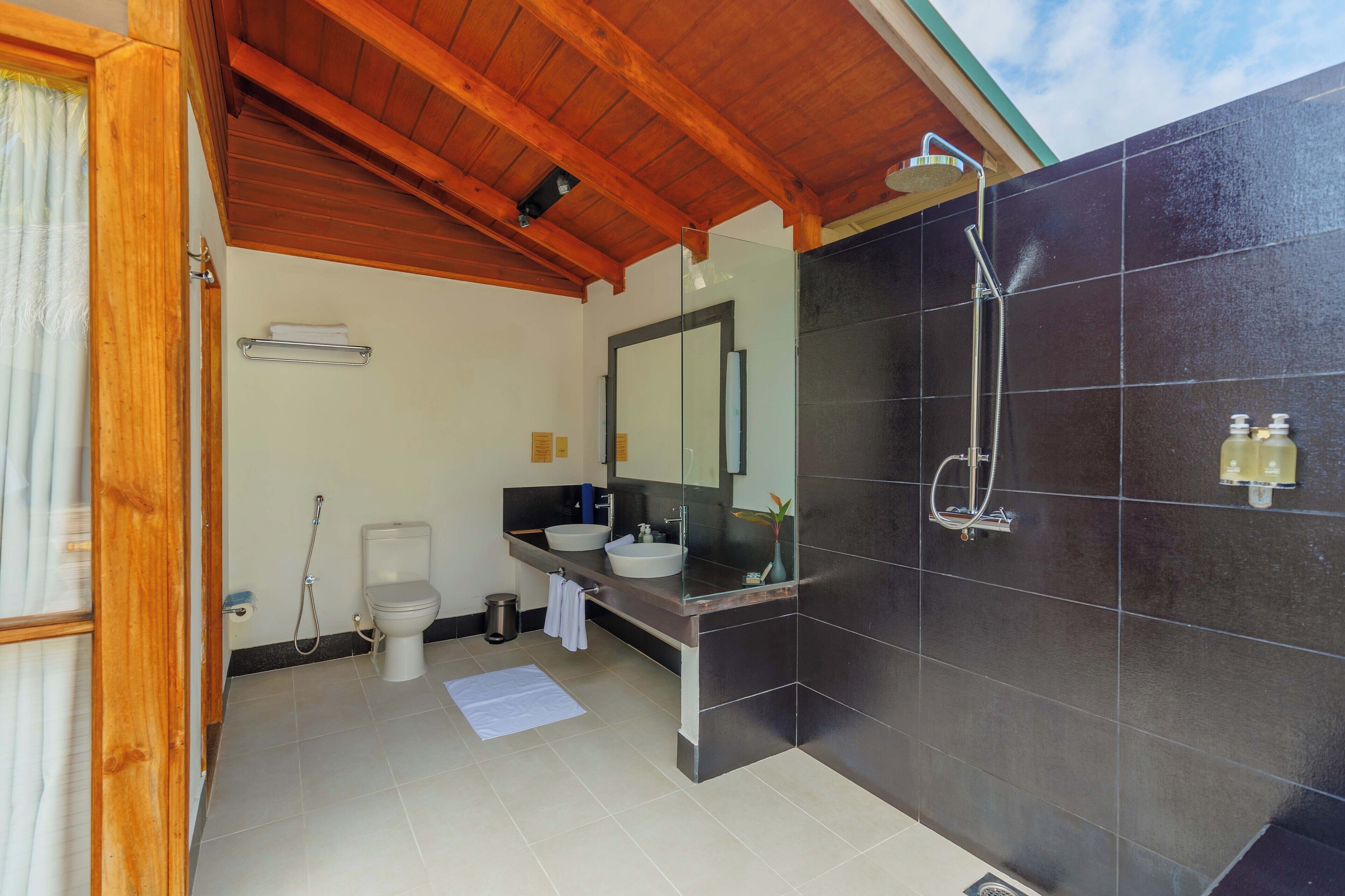 Une salle de bain moderne avec un plafond en bois, un grand miroir, une douche en verre et un mur recouvert de carreaux noirs.