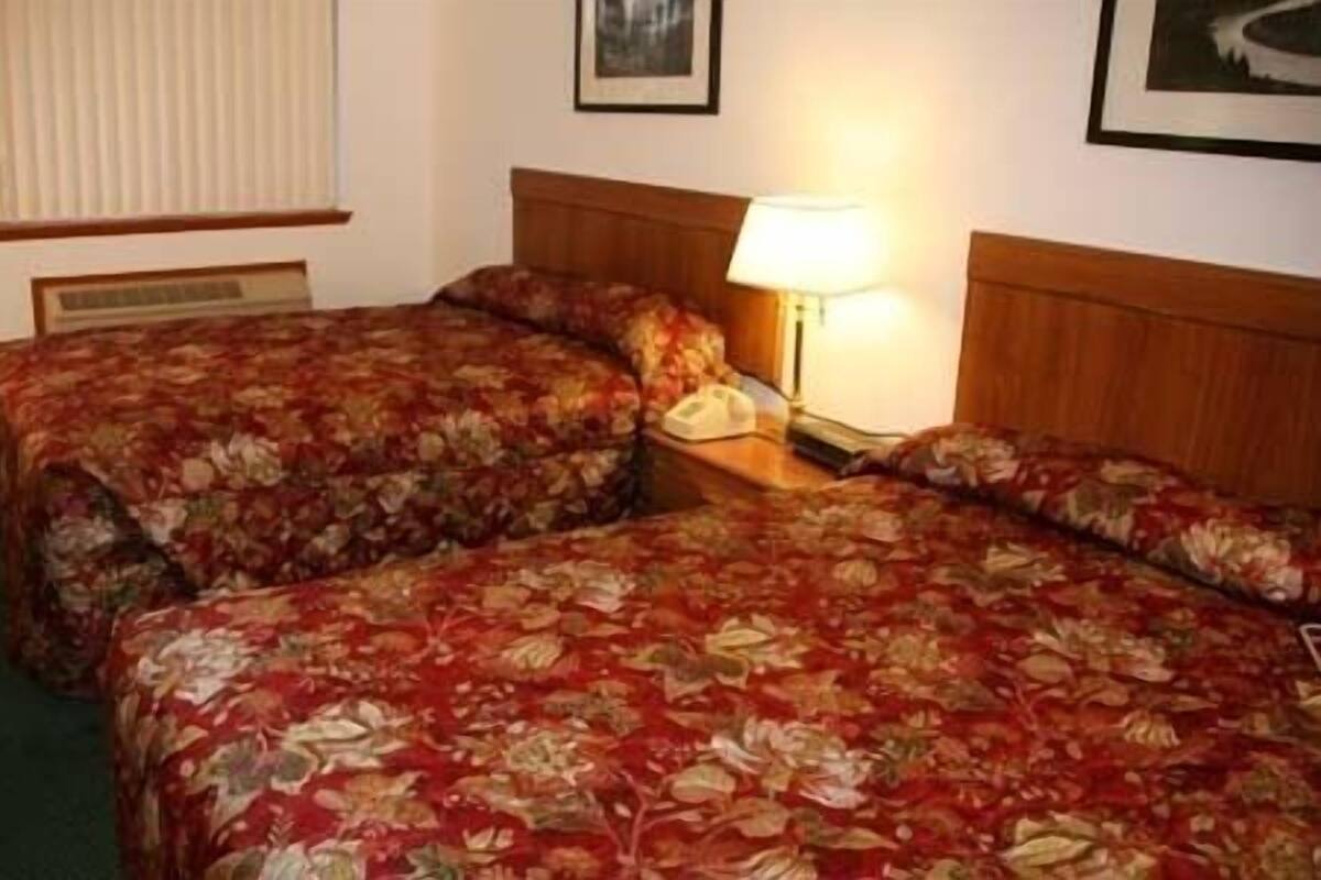 A hotel room with two beds, a bedside table with a lamp, and a window with blinds.