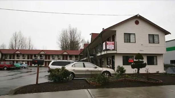 A motel with a two-story structure, a parking lot, and a red-roofed building in the background. - Sunshine Motel (Fife)