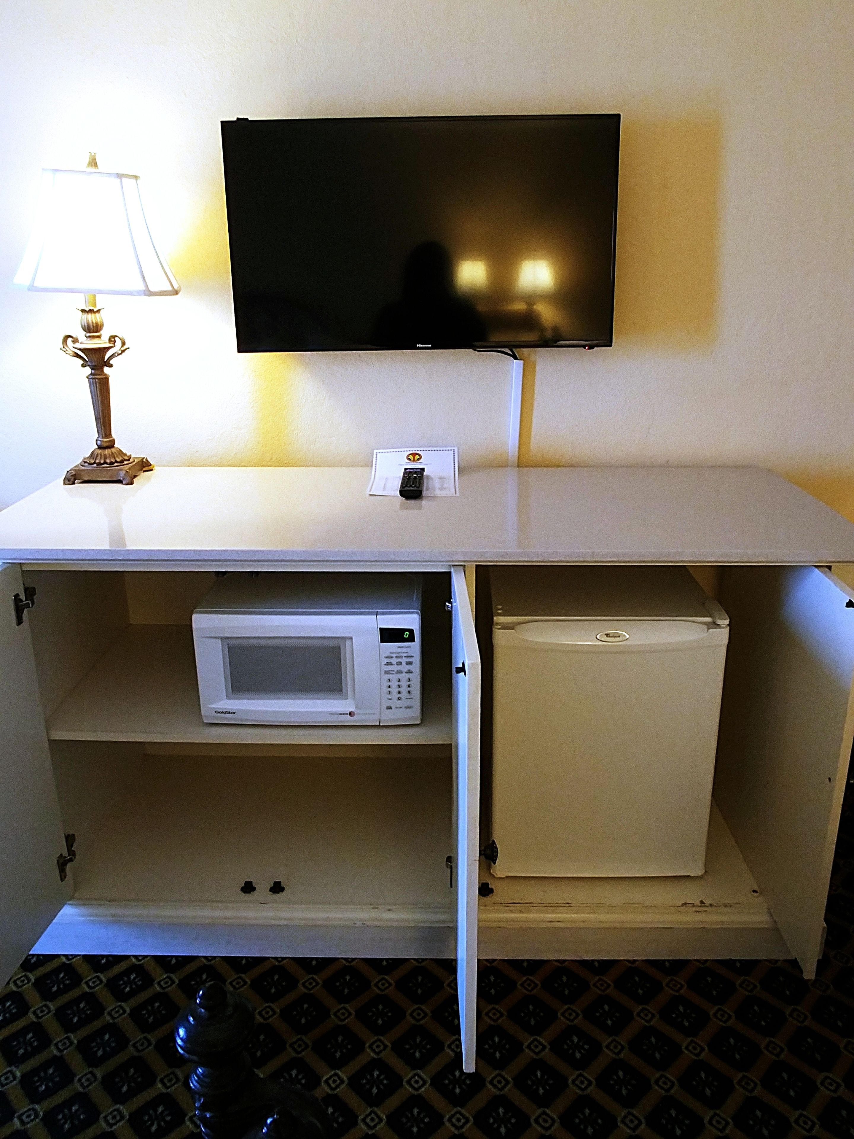A compact kitchenette with a microwave, a small refrigerator, a flat-screen TV mounted on the wall, and a table lamp.