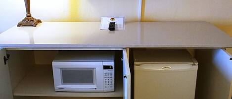 A compact kitchenette with a microwave, a small refrigerator, a flat-screen TV mounted on the wall, and a table lamp.