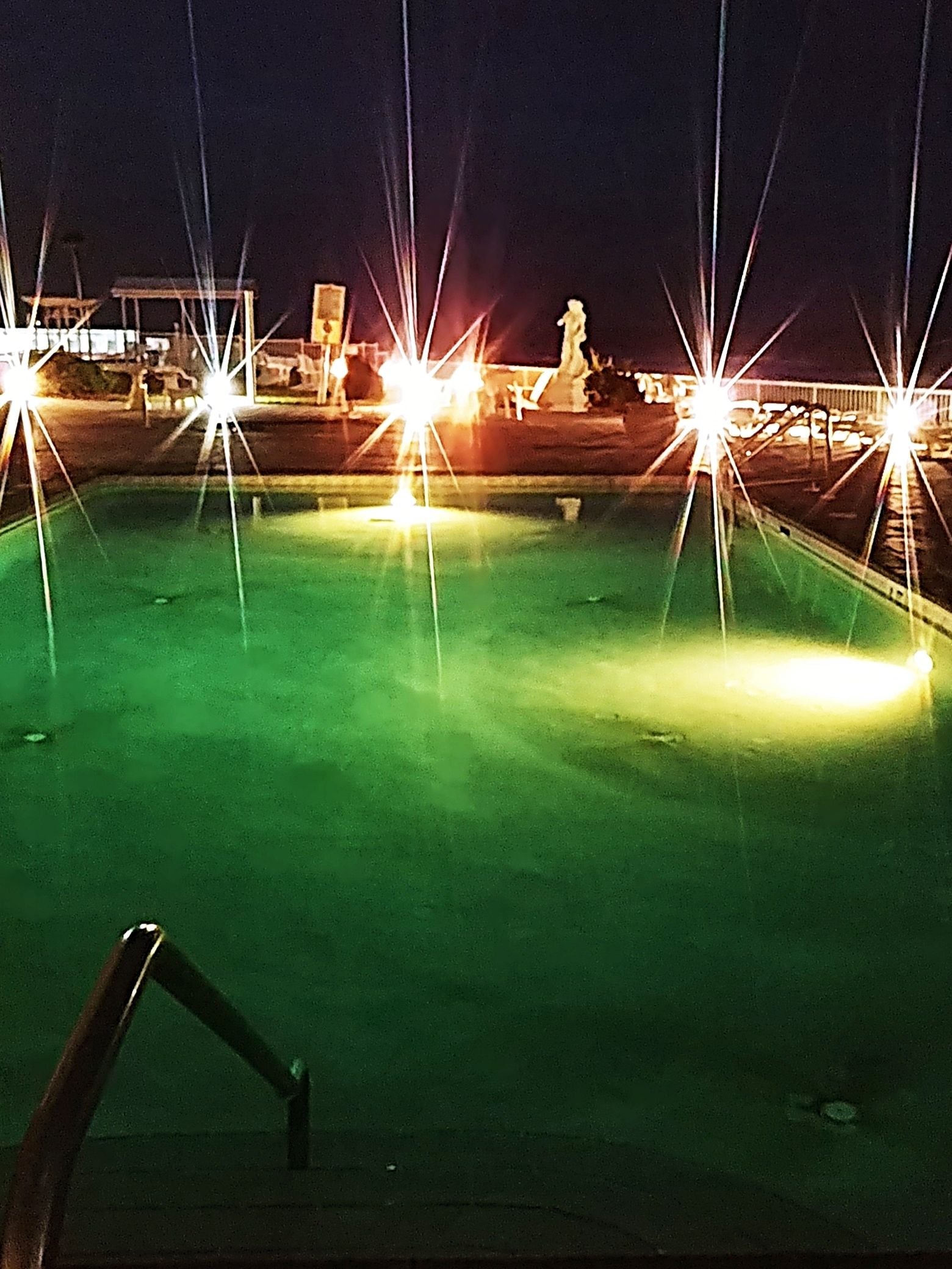 Vista nocturna de una piscina con luces brillantes y un trampolín visible.