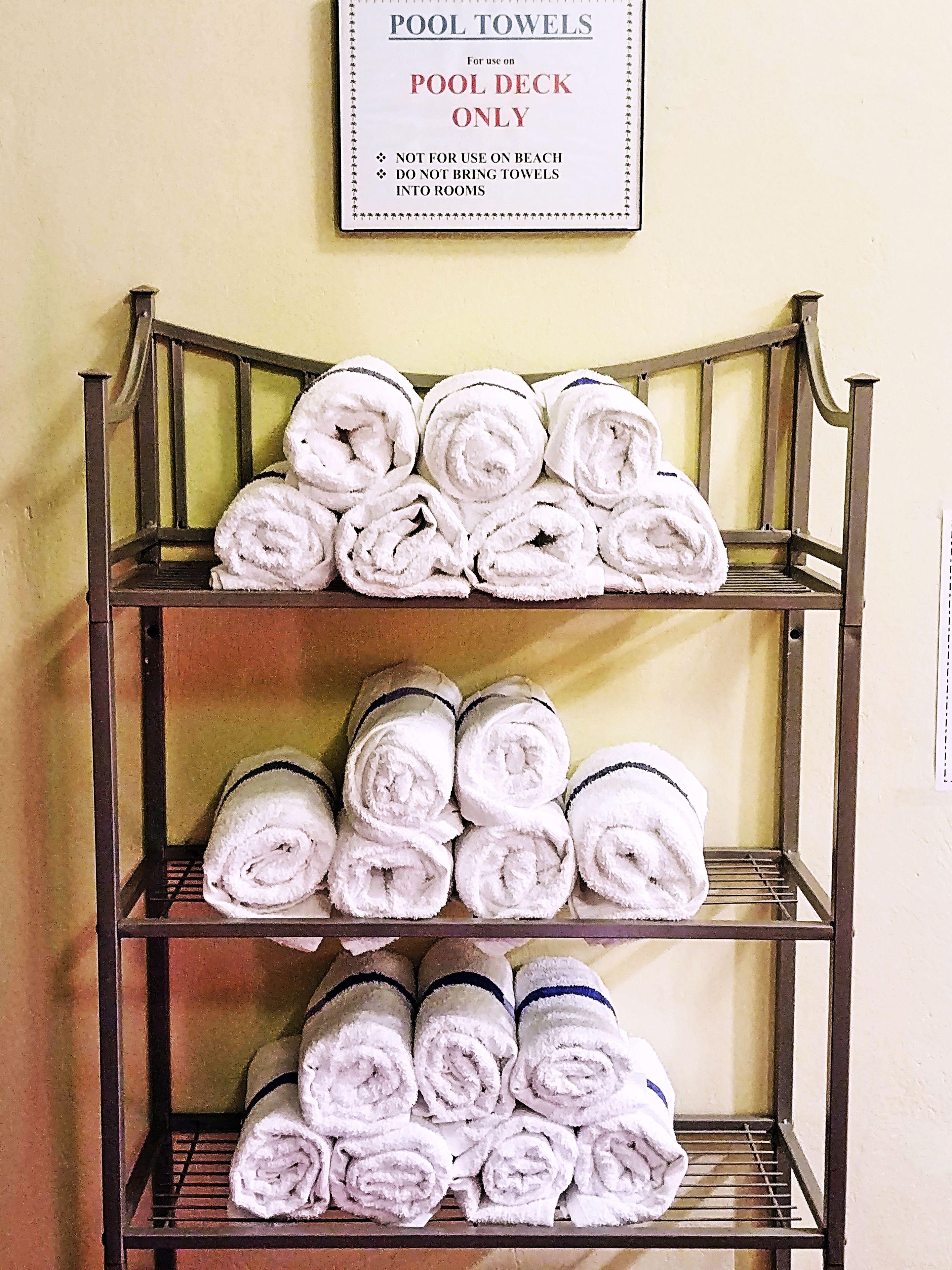 A metal shelf with neatly rolled white towels, a sign above stating 'POOL TOWELS For use on POOL DECK ONLY', and a wall with a light yellow hue.