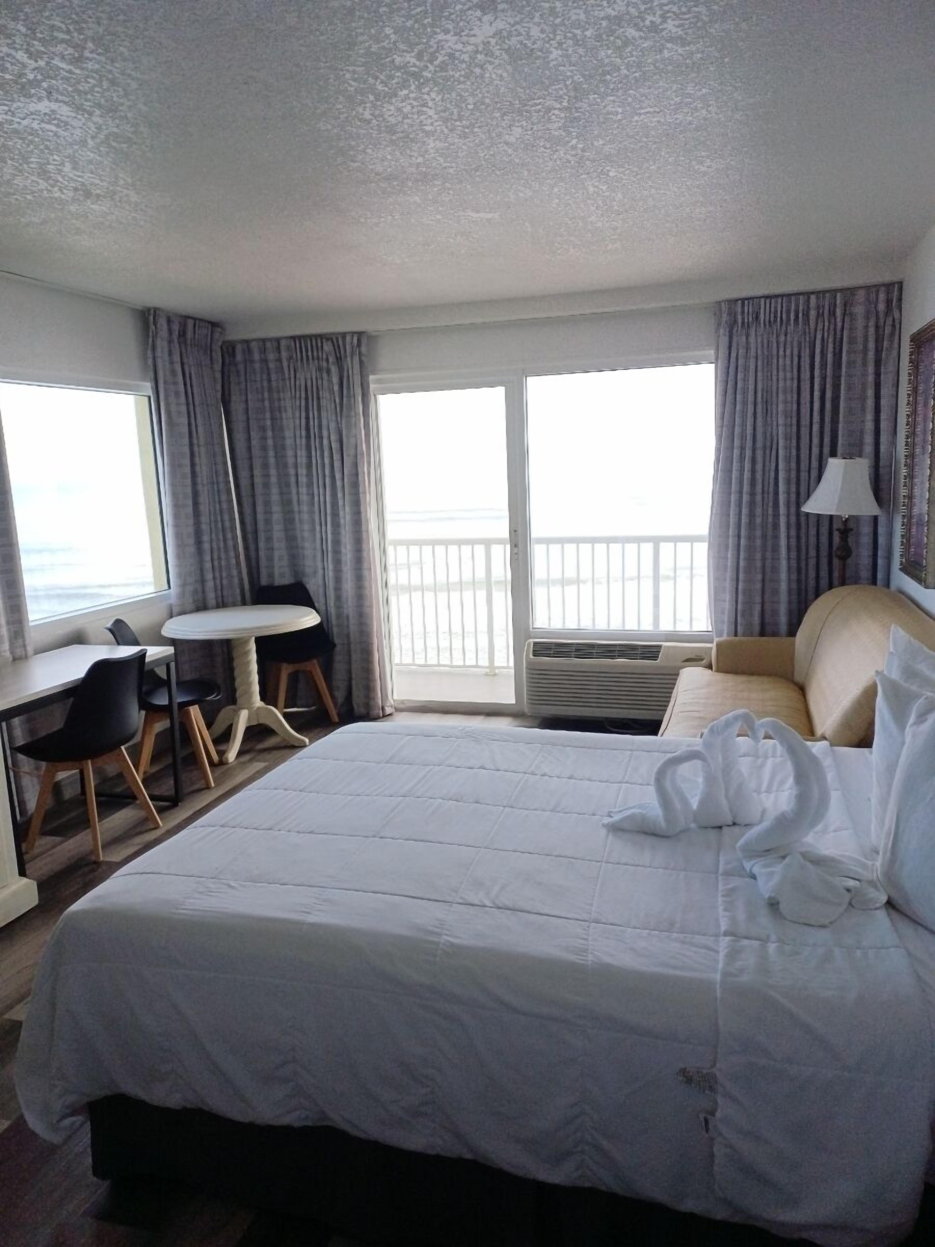 Habitación de hotel con una cama grande, un escritorio, dos sillas, un balcón con cortinas y una lámpara.