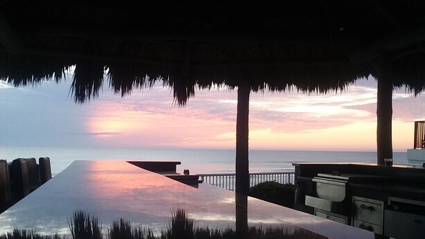 Vista a la playa con una estructura de techo de paja, una piscina y el atardecer sobre el océano.