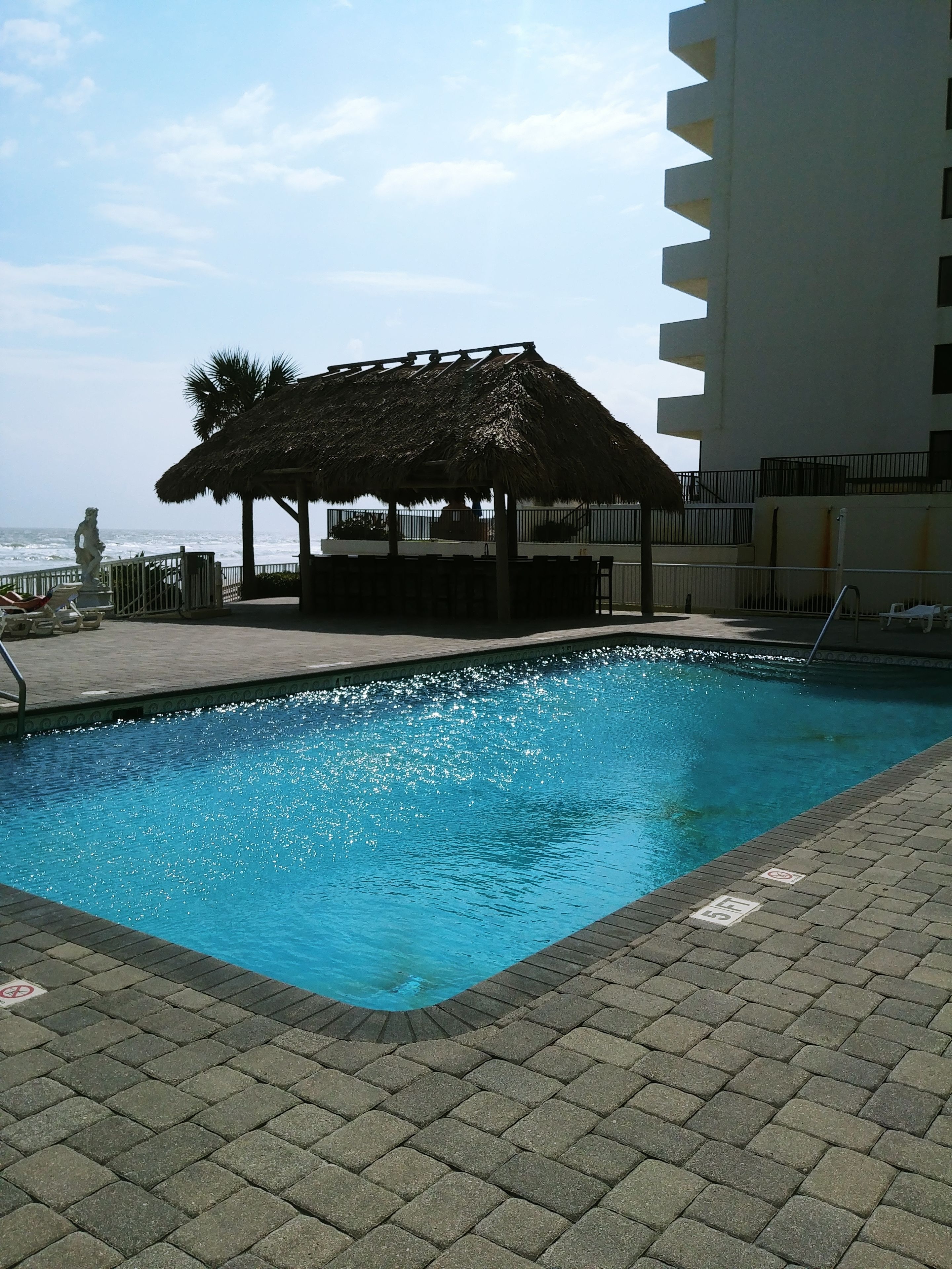 A swimming pool with a thatched-roof structure, a statue, and a view of the ocean.