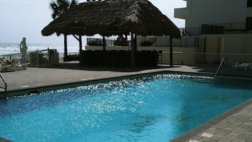 A swimming pool with a thatched-roof structure, a statue, and a view of the ocean.