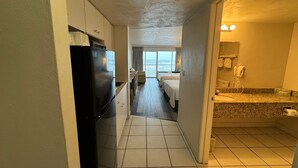 Un pasillo de hotel con una zona de cocina, un dormitorio con balcón y un baño con lavabo y espejo.