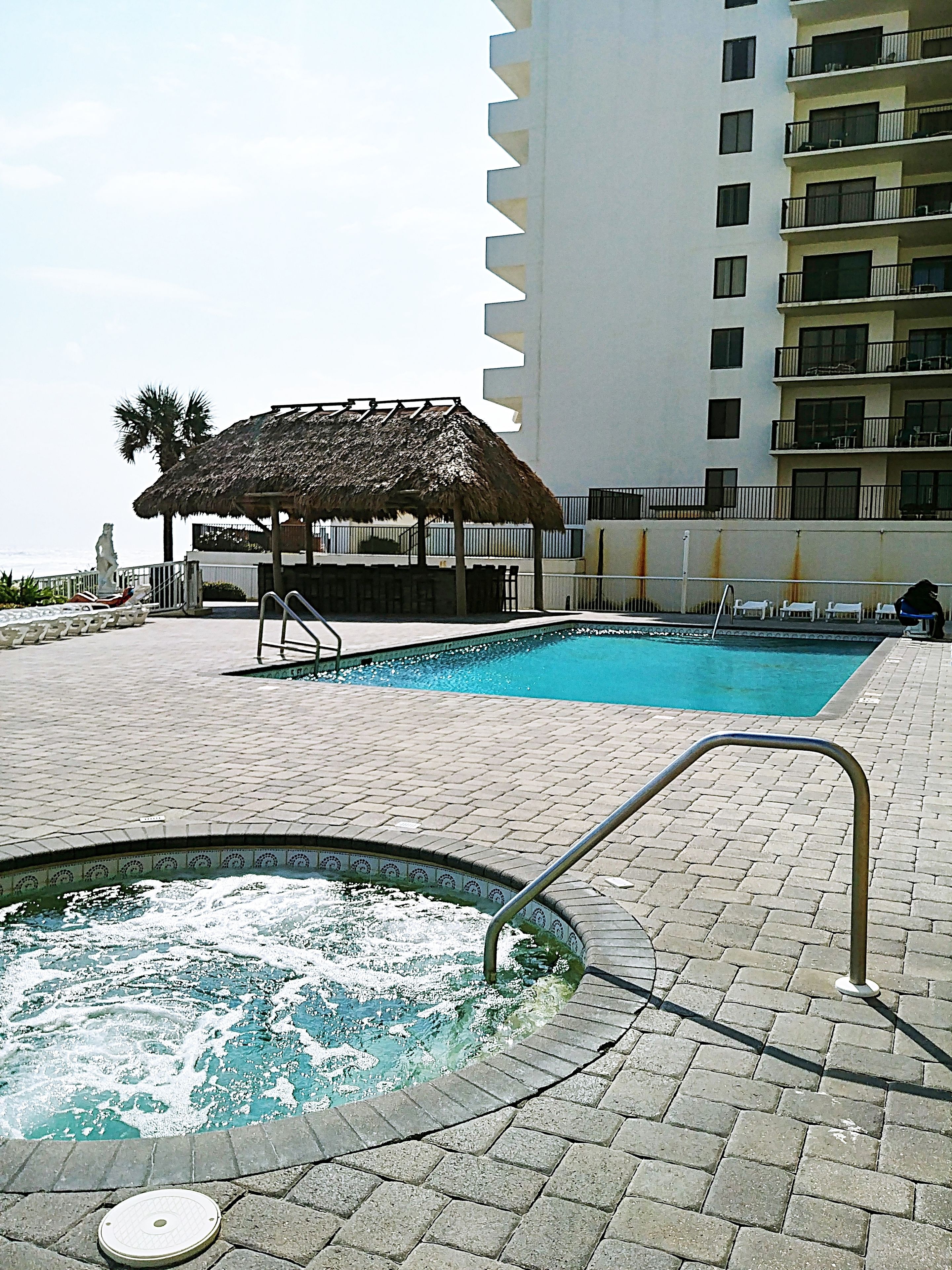 A swimming pool area with a hot tub, a thatched-roof gazebo, and a multi-story building in the background.