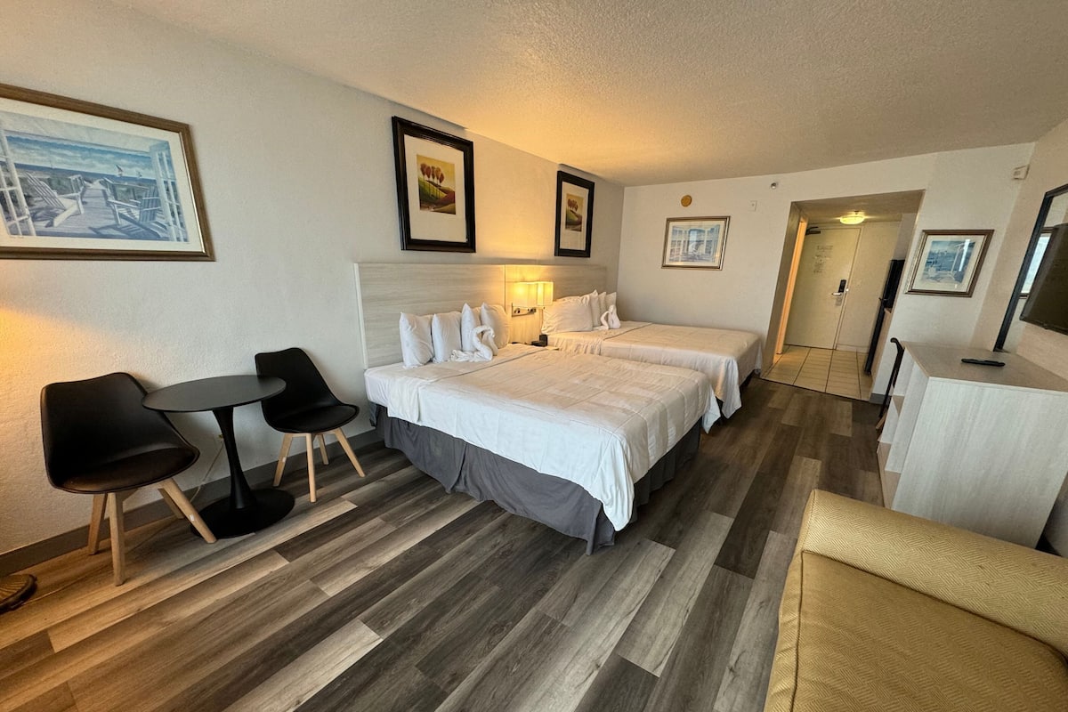 A hotel room with two beds, a desk, a chair, and a dining table.