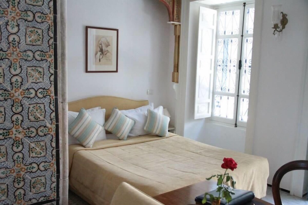 Un dormitorio con una cama, almohadas, una mesita de noche con un jarrón de flores, una silla y una ventana con cortinas.