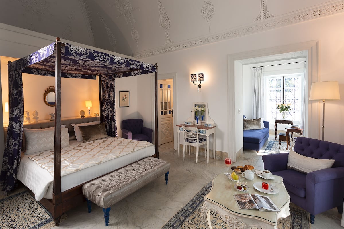 Una cama con dosel y cortinas, una zona de estar con sofá y sillones, una mesa de comedor con un jarrón de flores y una ventana con cortinas.