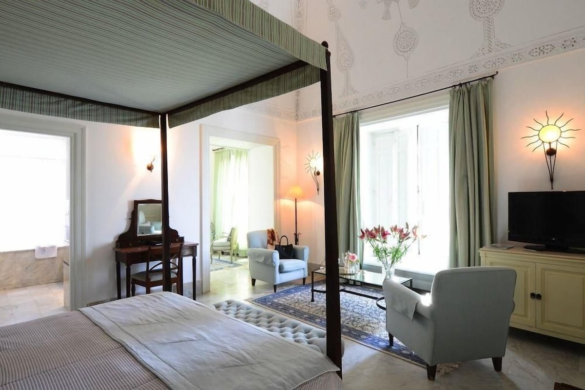 Un dormitorio con cama con dosel, un escritorio con espejo, un televisor y una zona de estar con una silla y una mesa.
