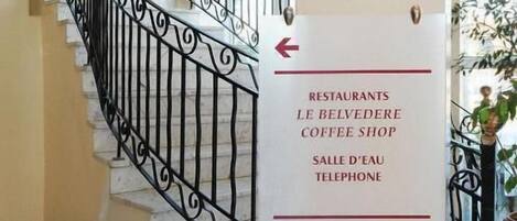 Interior de un hotel con una escalera, un panel indicando el camino a restaurantes, una cafetería y habitaciones 101 a 123.