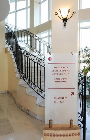 A hotel interior with a staircase, a signpost indicating the direction to restaurants, a coffee shop, and rooms 101-123. - Hotel du Parc (Tunis)