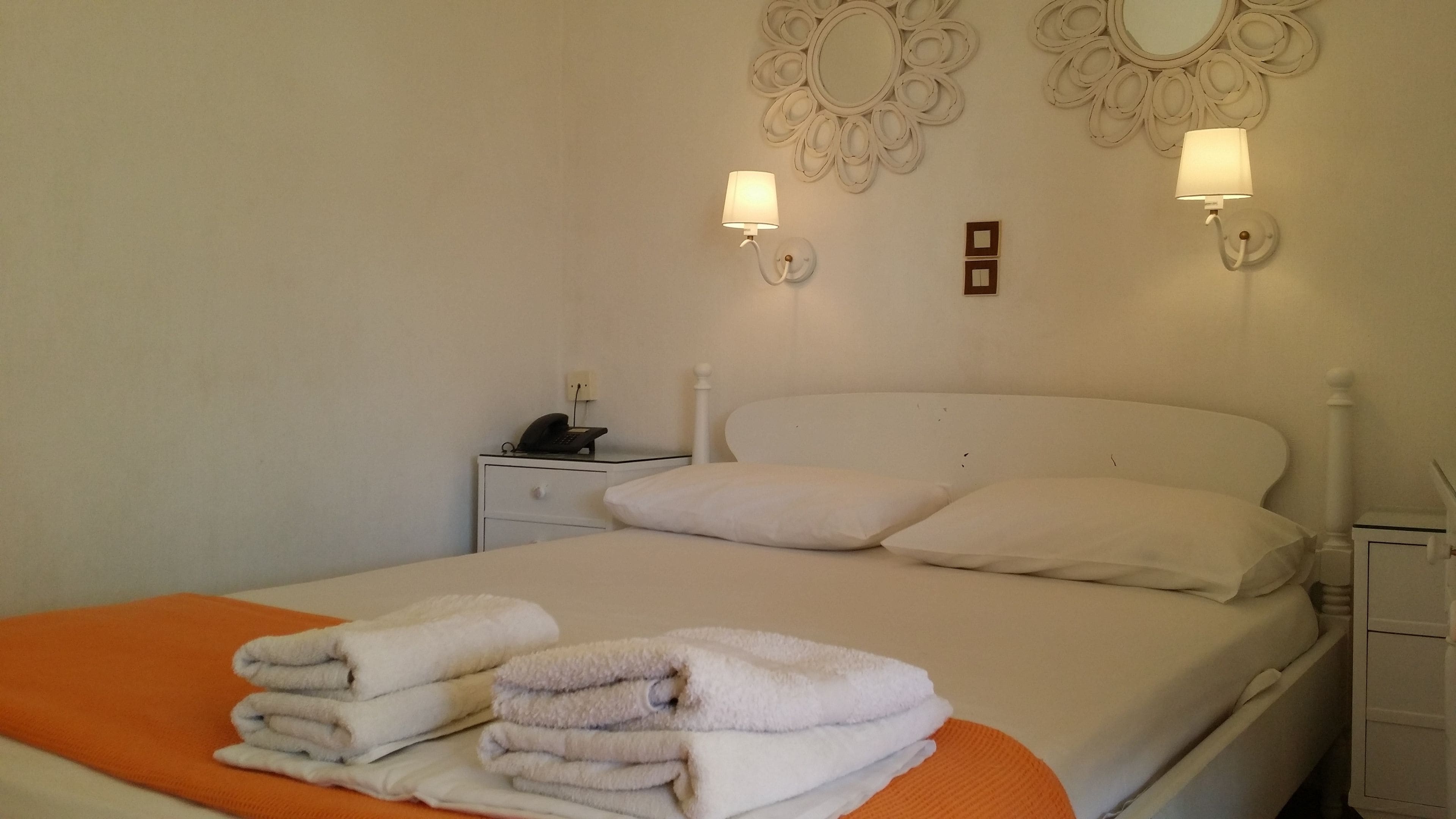 Un lit bien fait, avec du linge de lit blanc et une couverture orange, une table de chevet avec un téléphone et deux lampes fixées au mur.