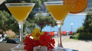 Two martini glasses with a yellow liquid, an orange slice, and a candle on a table with a scenic view of a beach and trees in the background. - Hotel Rachel (Aegina)