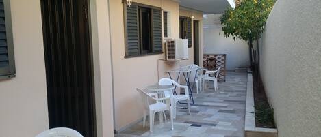 A small outdoor patio with white plastic chairs and tables, air conditioning units, and a small garden area with a tree.