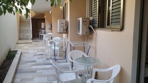 An outdoor seating area with white plastic chairs and tables, air conditioning units mounted on the wall, and a tiled floor. - Hotel Rachel (Aegina)