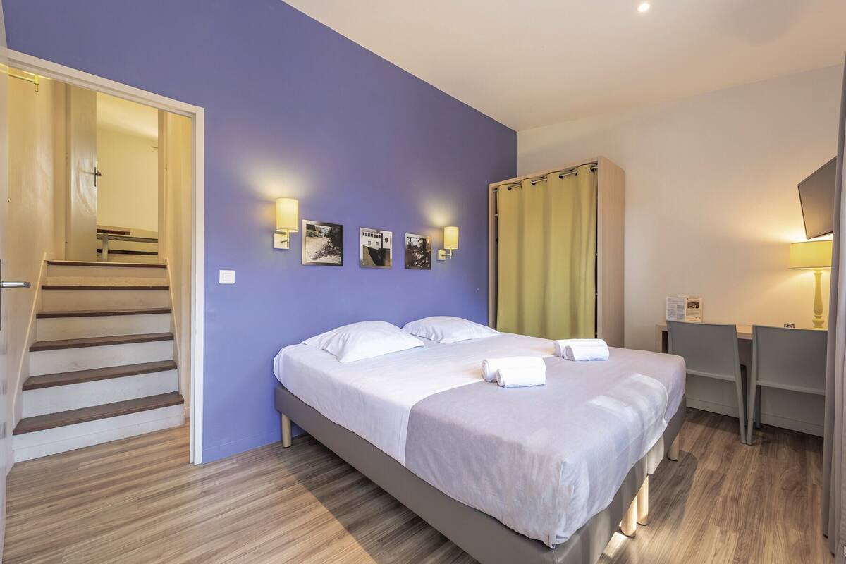 Un dormitorio con cama, escritorio y silla. Se ve una escalera que lleva a otra habitación.