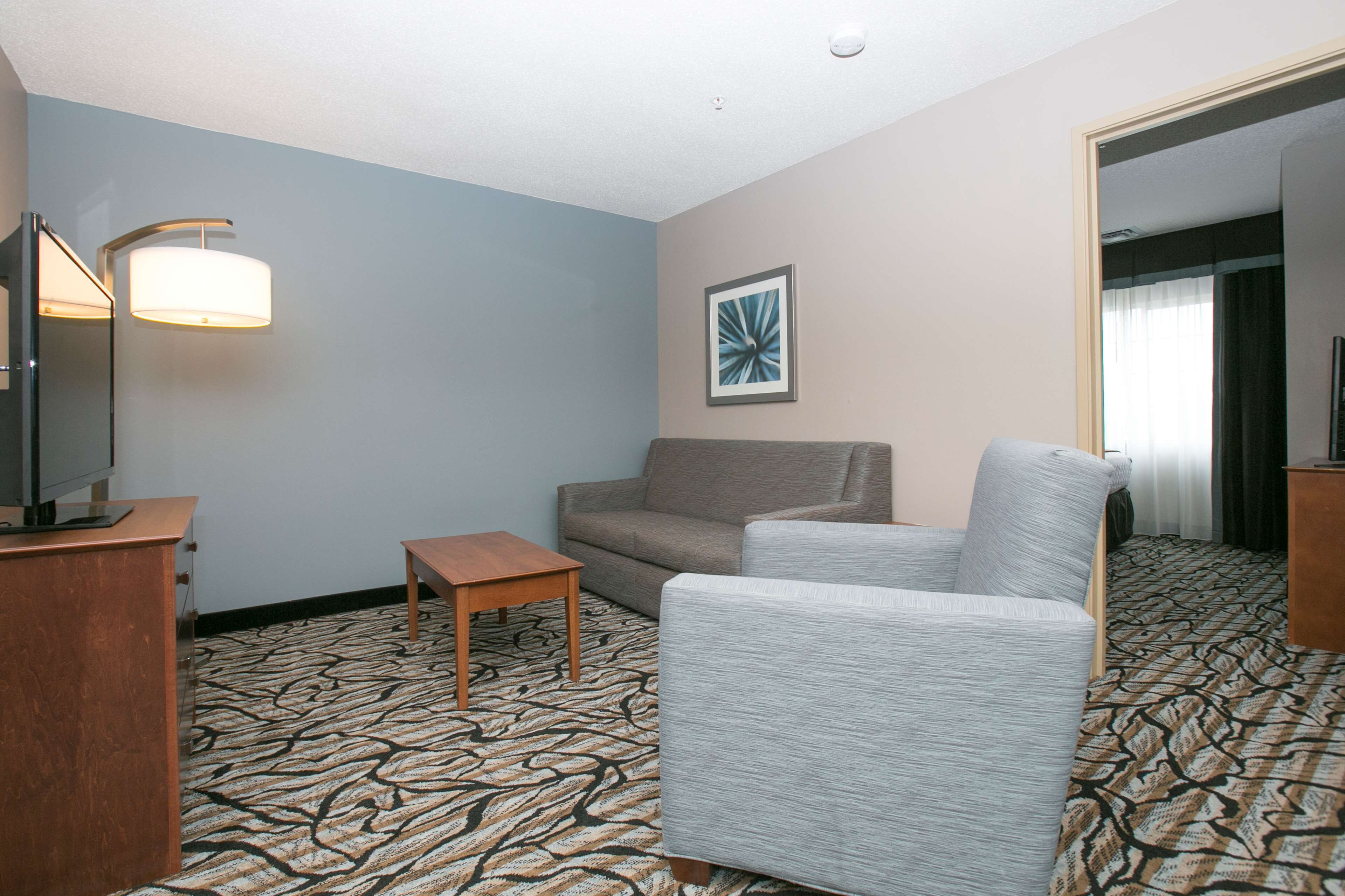 A hotel room with a patterned carpet, a grey sofa, a small wooden table, a television on a wooden stand, and a framed picture on the wall.