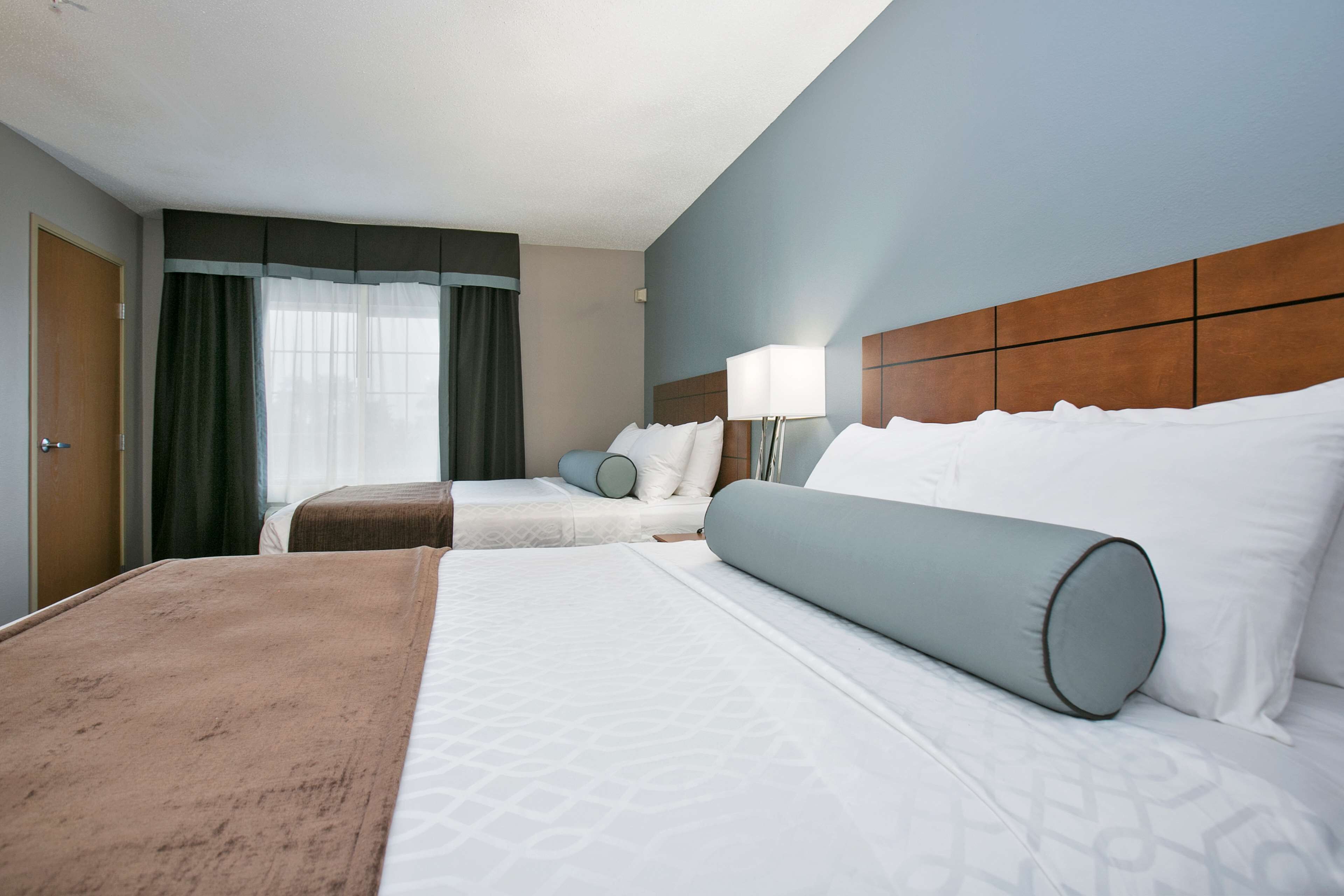 A hotel room with two beds, a large window with curtains, a wooden headboard, and a bedside lamp.