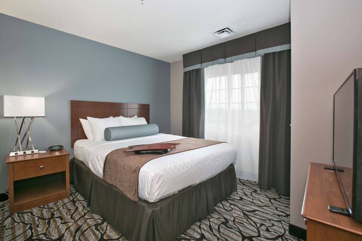 A hotel room with a bed, a nightstand, a lamp, a television, and a window with curtains.
