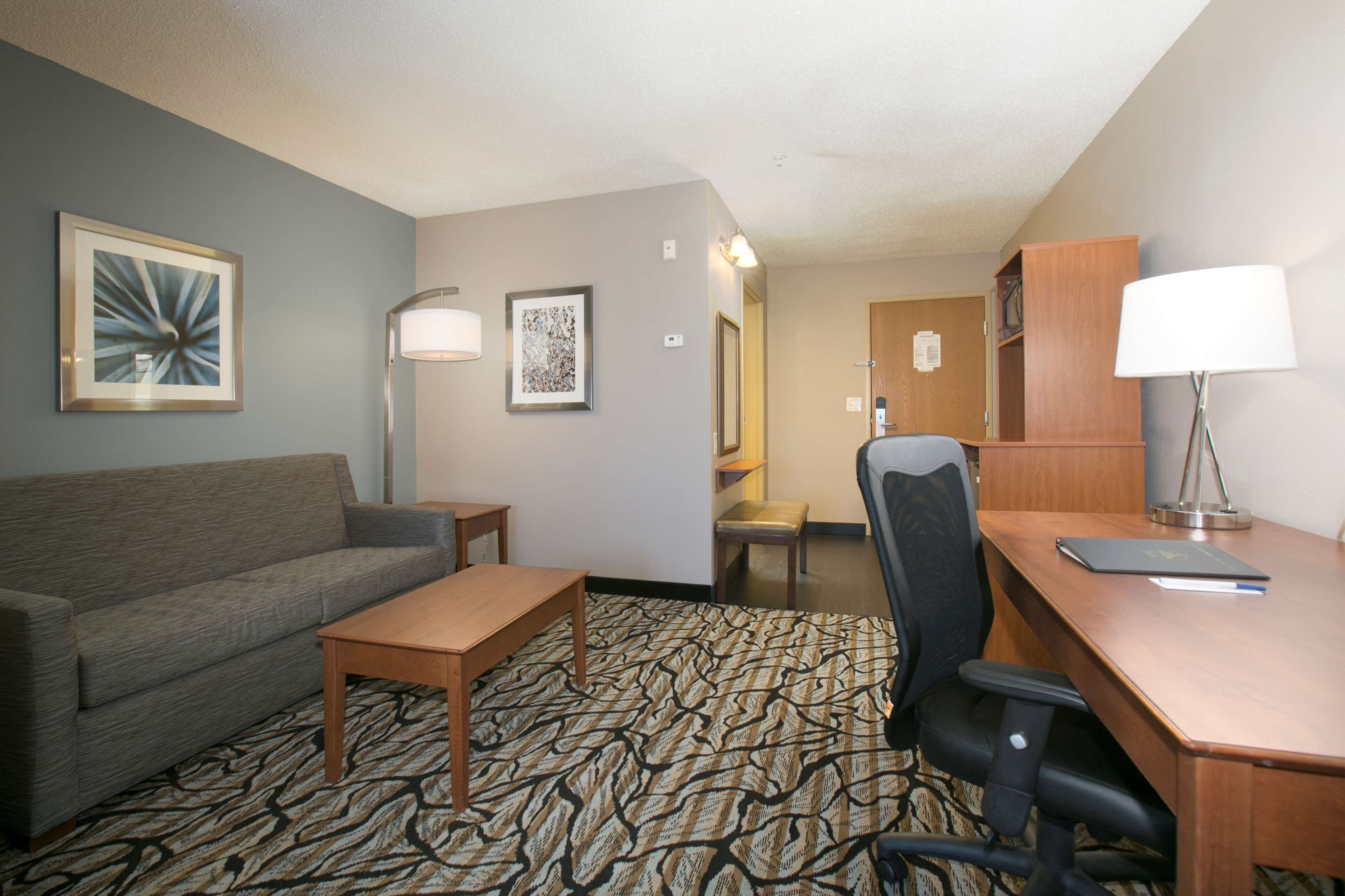 A hotel room with a sofa, a small table, a desk with a chair, a patterned carpet, and framed artwork on the walls.