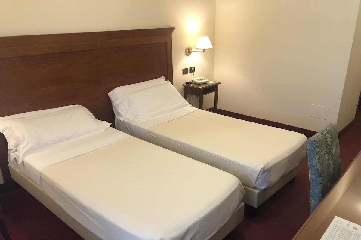 Two single beds with white linens, a wooden headboard, a bedside table with a lamp, and a small desk with a chair.