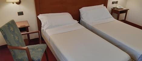 Habitación de hotel con dos camas individuales, cabecero de madera, una silla, una mesita de noche y una lámpara.