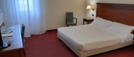 A hotel room with a bed, a desk, a chair, a nightstand, and a lamp.