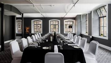 Un salón de conferencias con una larga mesa, sillas blancas y ventanas amplias con vistas a edificios.