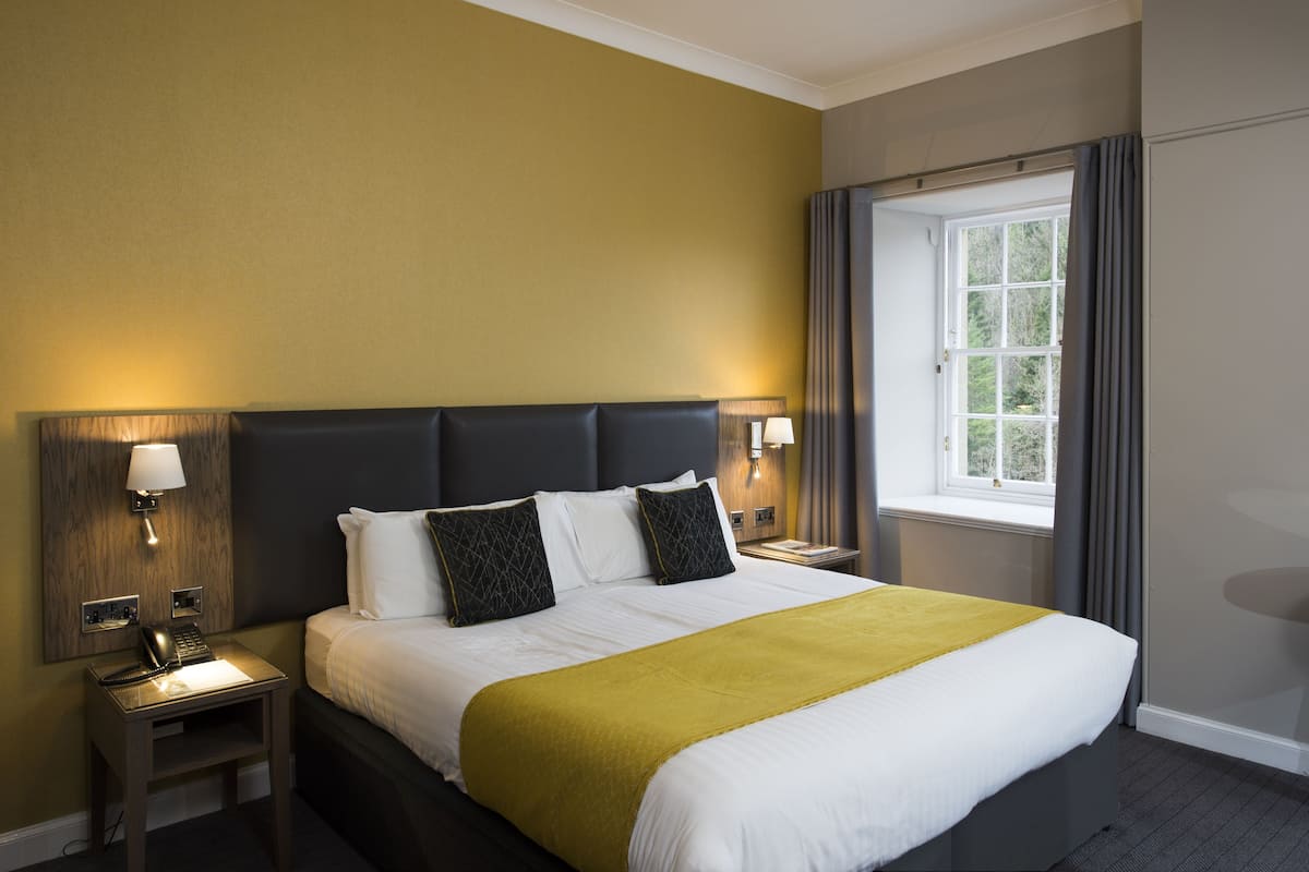 A hotel room with a large bed, two bedside tables with lamps, a window with curtains, and a view of greenery outside.