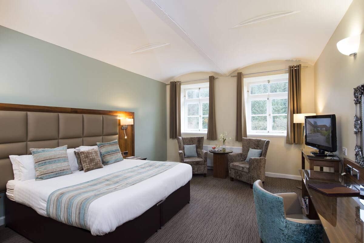 A hotel room with a large bed, two armchairs, a desk with a TV, and a window with curtains.