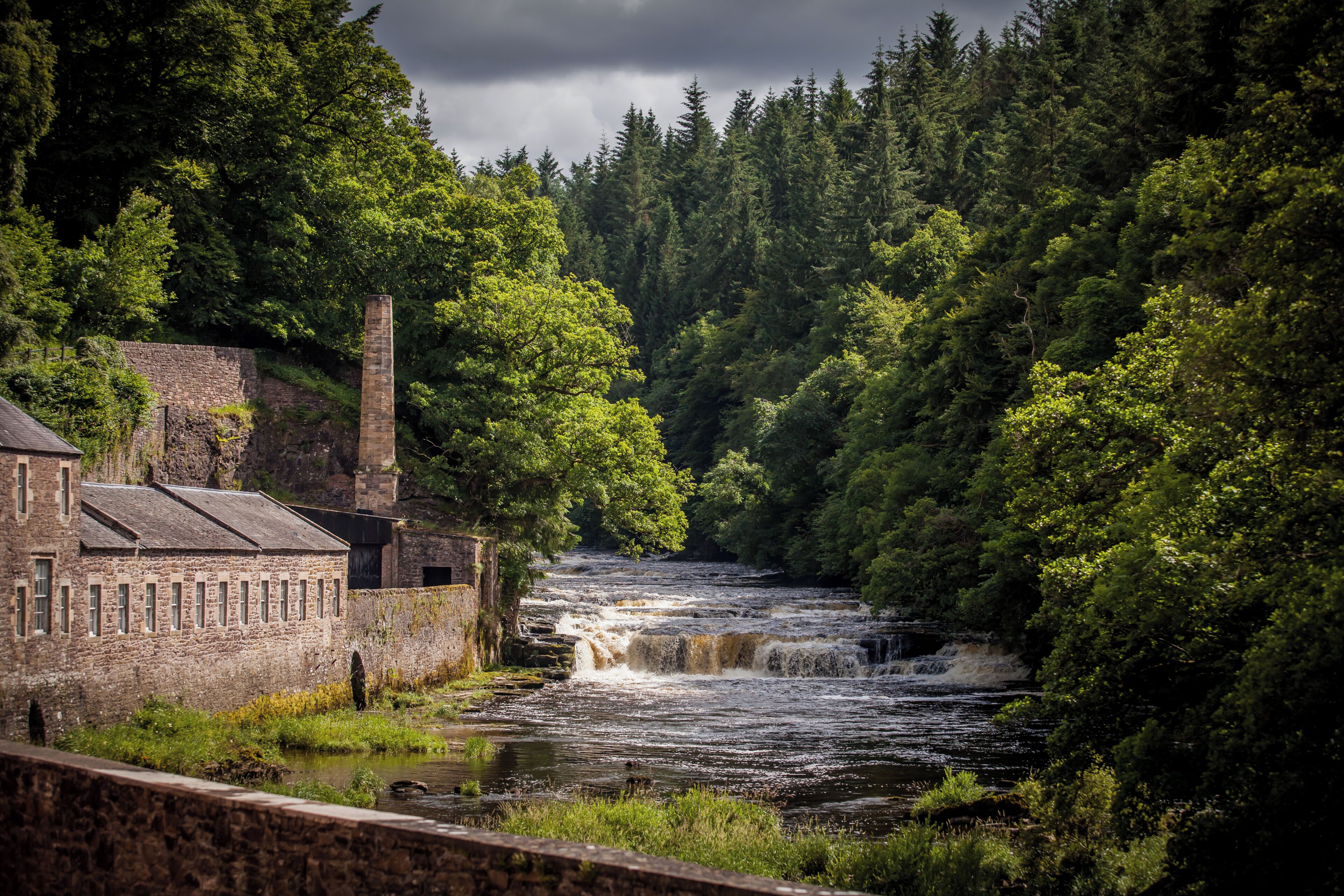 A river with a waterfall, surrounded by dense forest and a stone building with multiple windows.