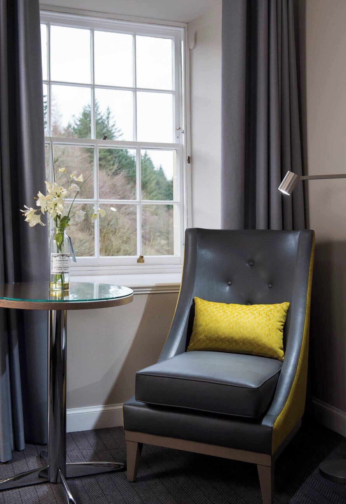 A room with a window, a chair with a yellow cushion, a glass table with a bottle, and a lamp.