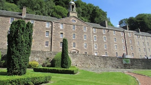 A large stone building with multiple windows and a central tower, surrounded by well-maintained gardens and trees. - New Lanark Mill Hotel (Lanark)