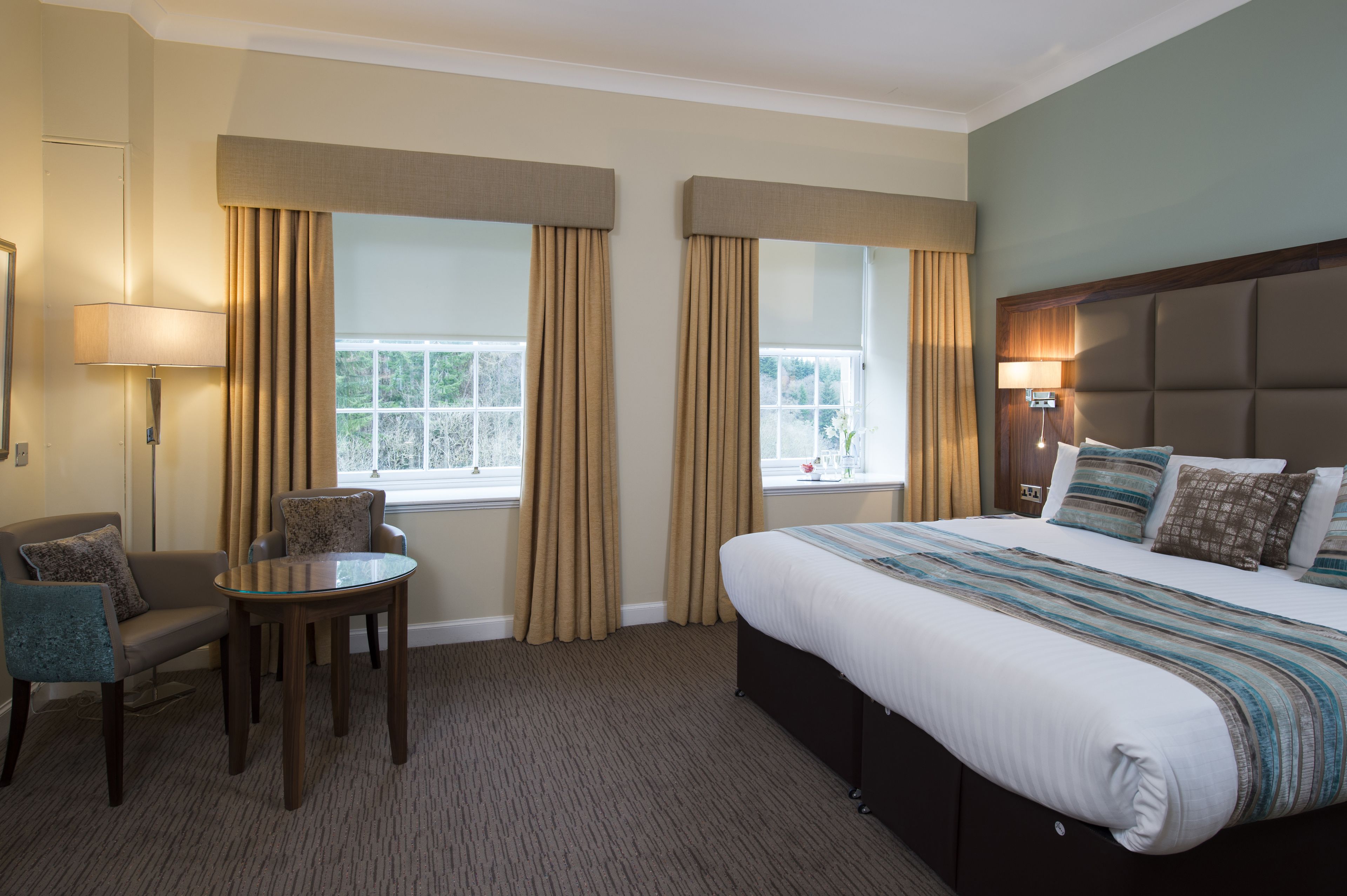 A hotel room with a large bed, a small table, a chair, and two windows with curtains.