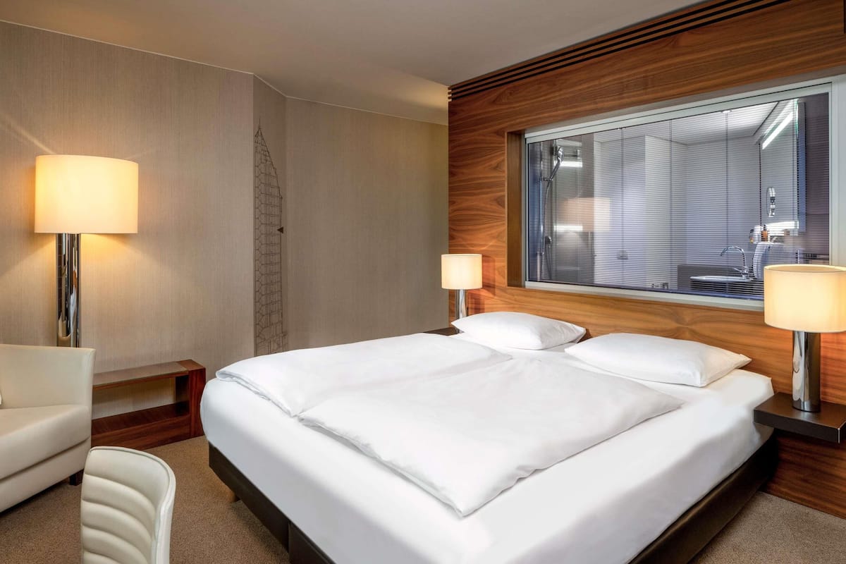 A hotel room with a large bed, two bedside lamps, a wooden headboard, a window with blinds, and a chair.