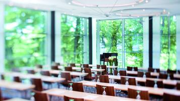 A conference room with rows of tables and chairs, large windows with a view of greenery, and a modern ceiling design.