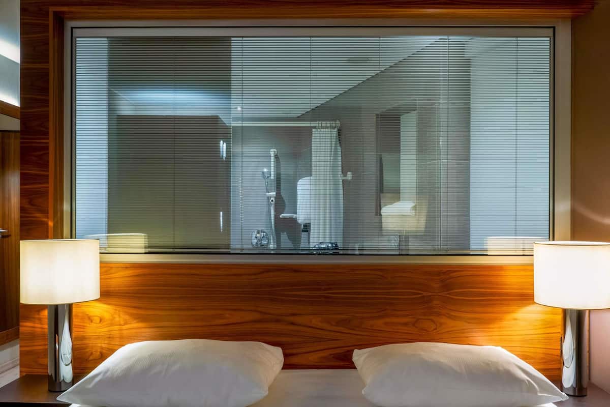 A hotel room with a large window, wooden headboard, two bedside lamps, and two pillows.