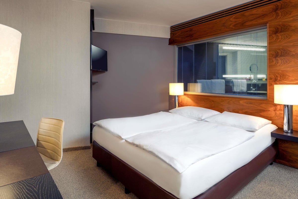 A hotel room with a bed, a TV mounted on the wall, a window with blinds, and a desk with a chair.