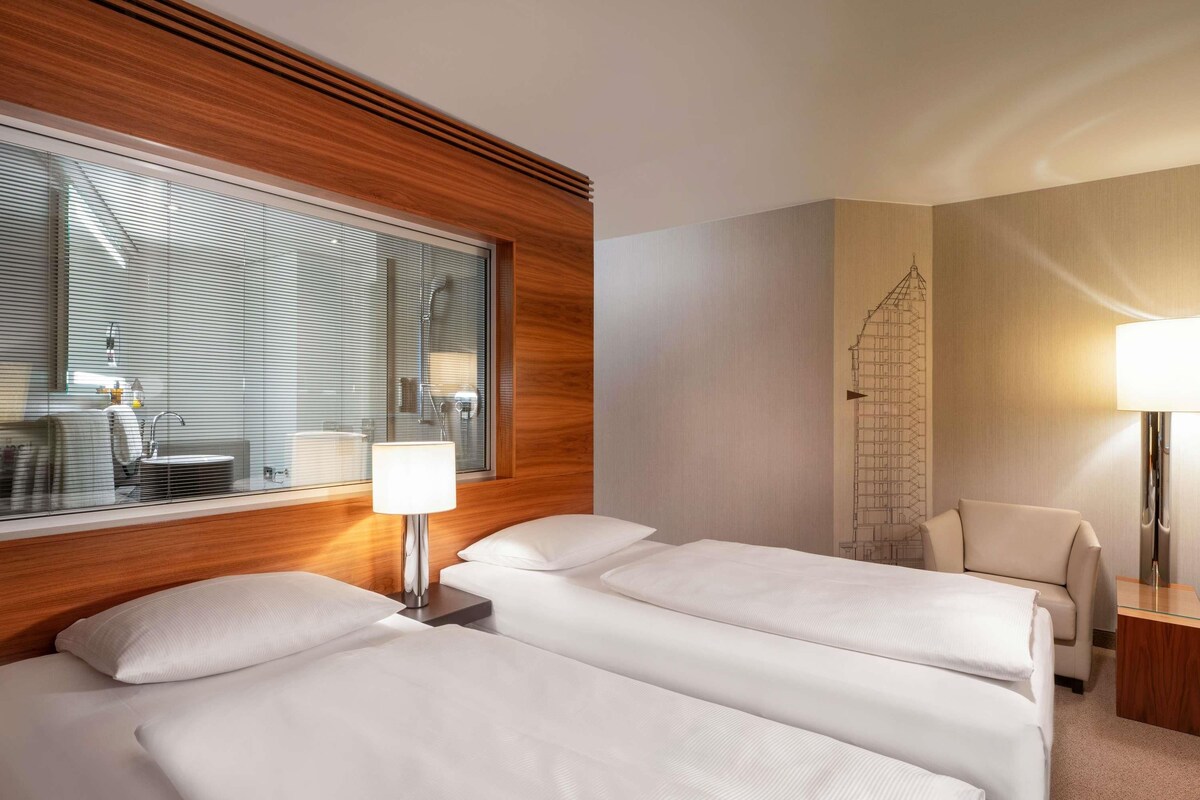 A hotel room with two beds, a wooden headboard, a large window with blinds, a desk, and a chair.