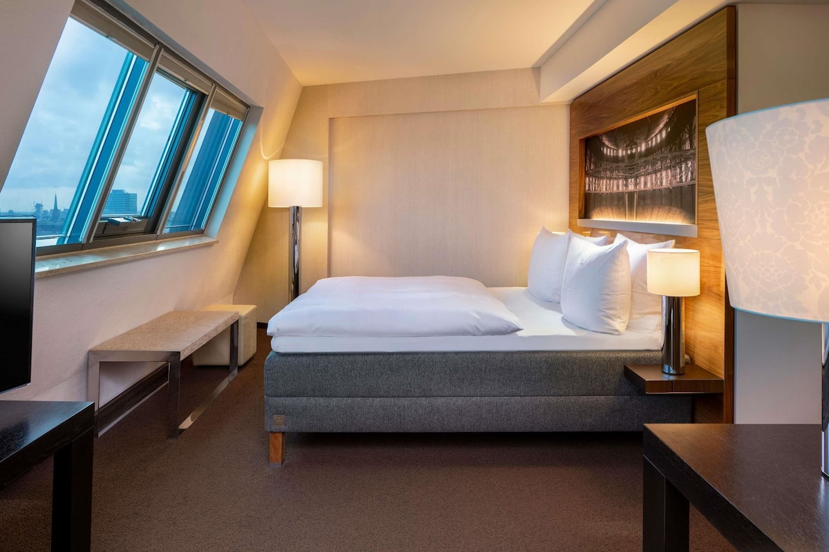 A modern hotel room with a large bed, two bedside lamps, a bench, a TV, and a city view through skylights.