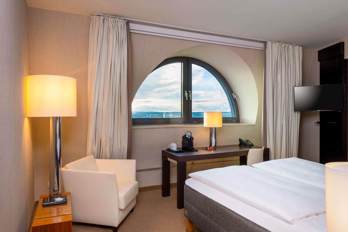 A hotel room with a bed, a desk, a chair, a lamp, a TV, and a window with a view.