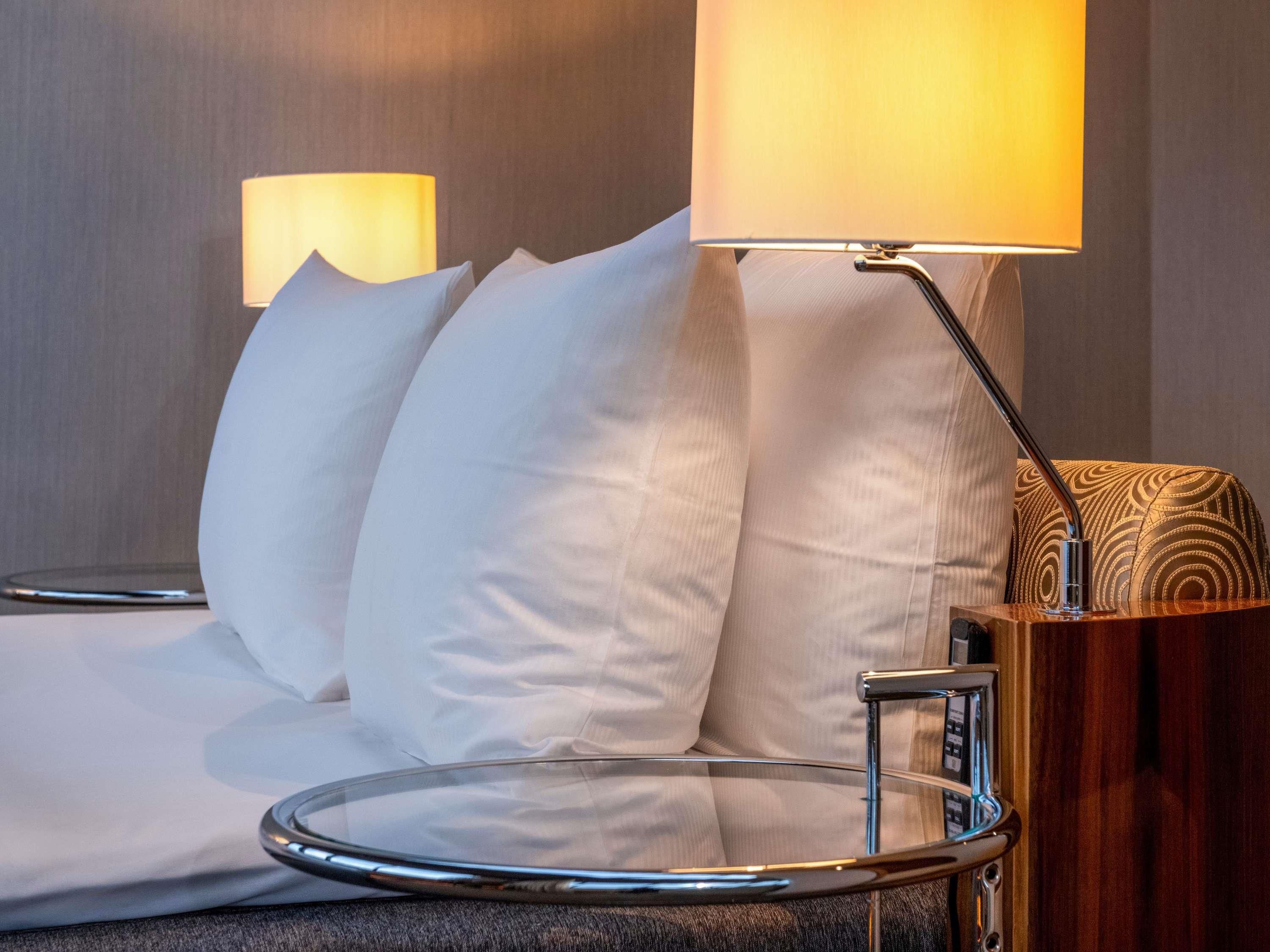 A modern hotel room with a sofa, a side table with a lamp, and a wooden side table with a patterned cushion.