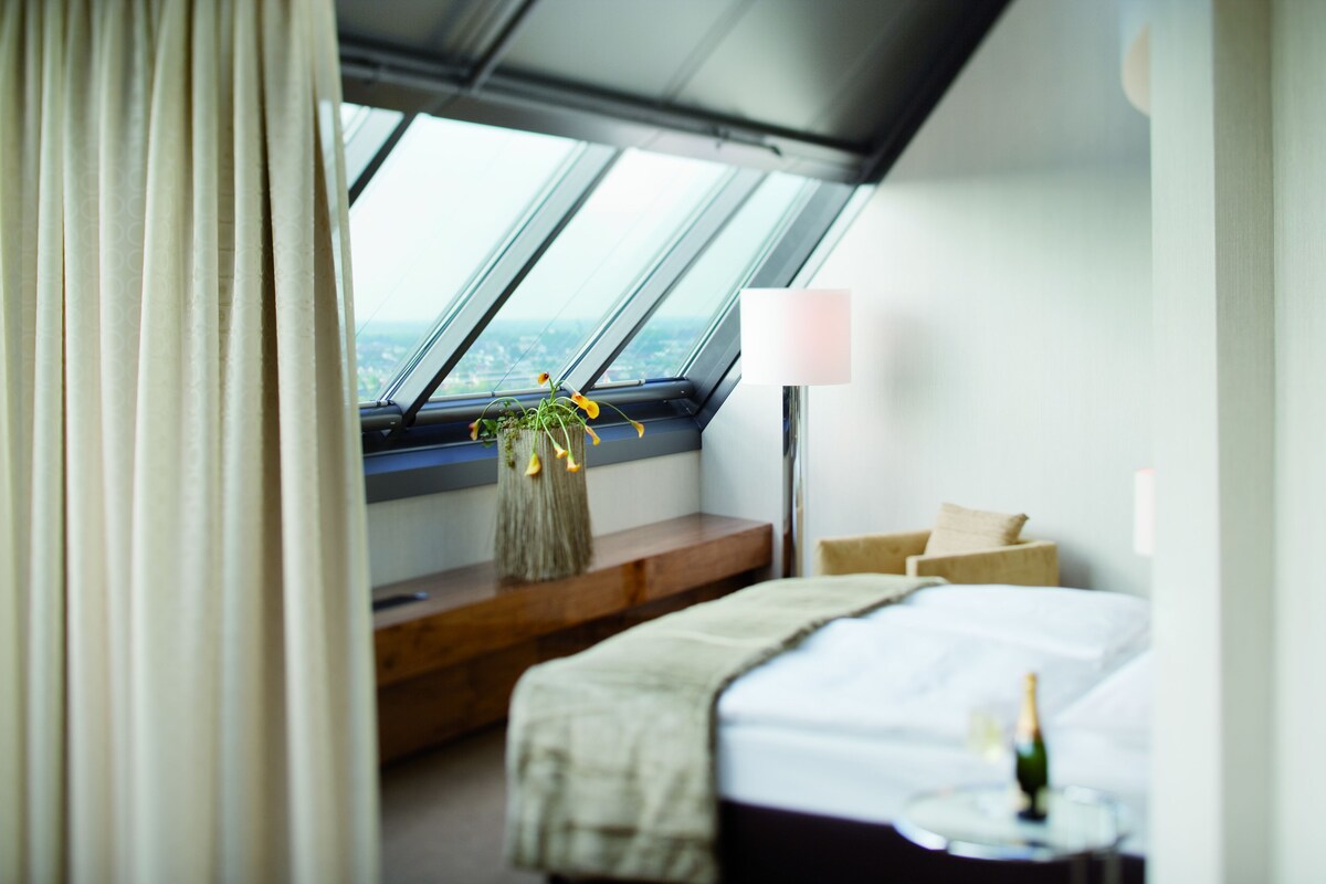 Ein modernes Hotelzimmer mit einem großen Dachfenster, einem Bett, einem Nachttisch mit einer Flasche und Blick auf die Stadt.