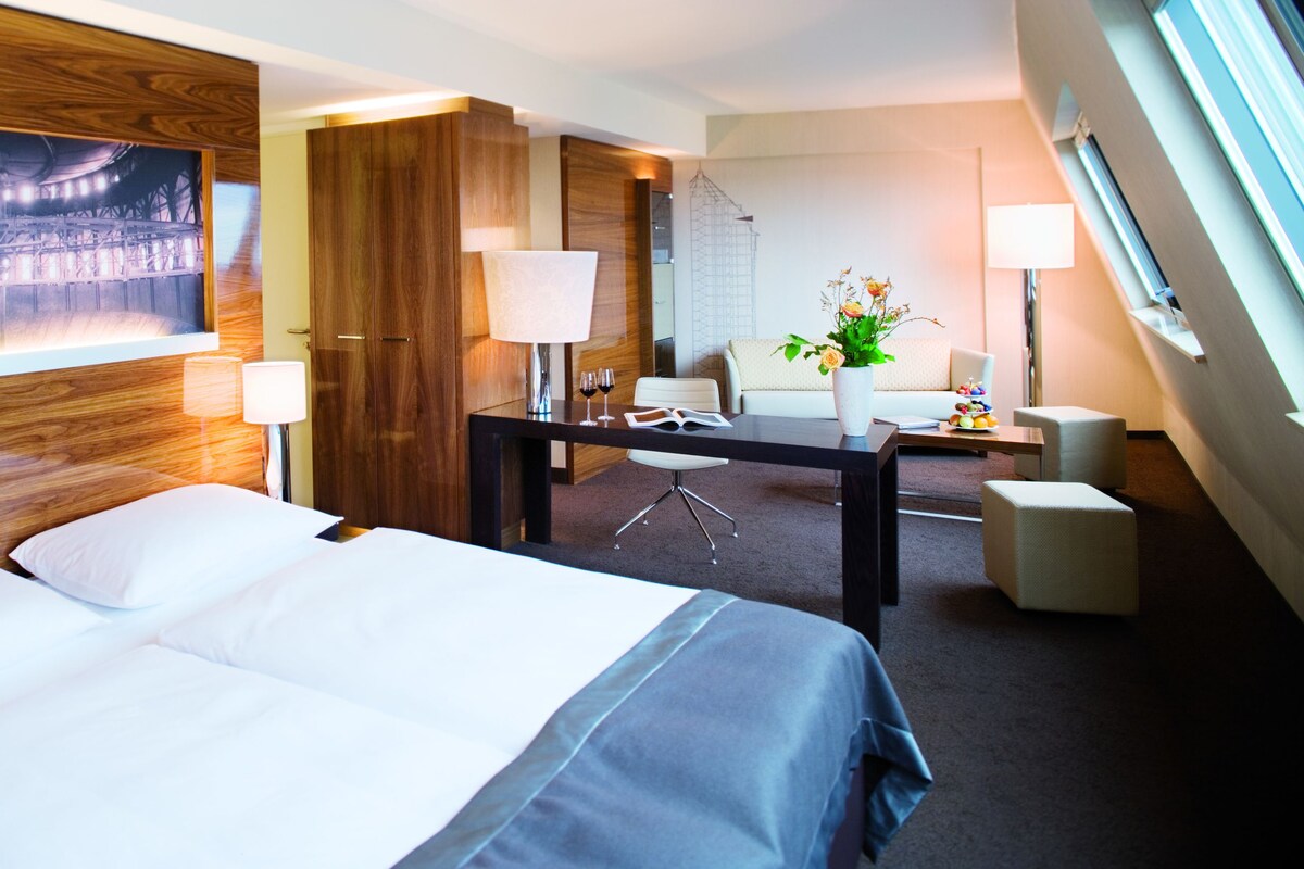 Ein Hotelzimmer mit einem großen Bett, einem Schreibtisch mit Stuhl, einer Lampe und einem großen Gemälde an der Wand.