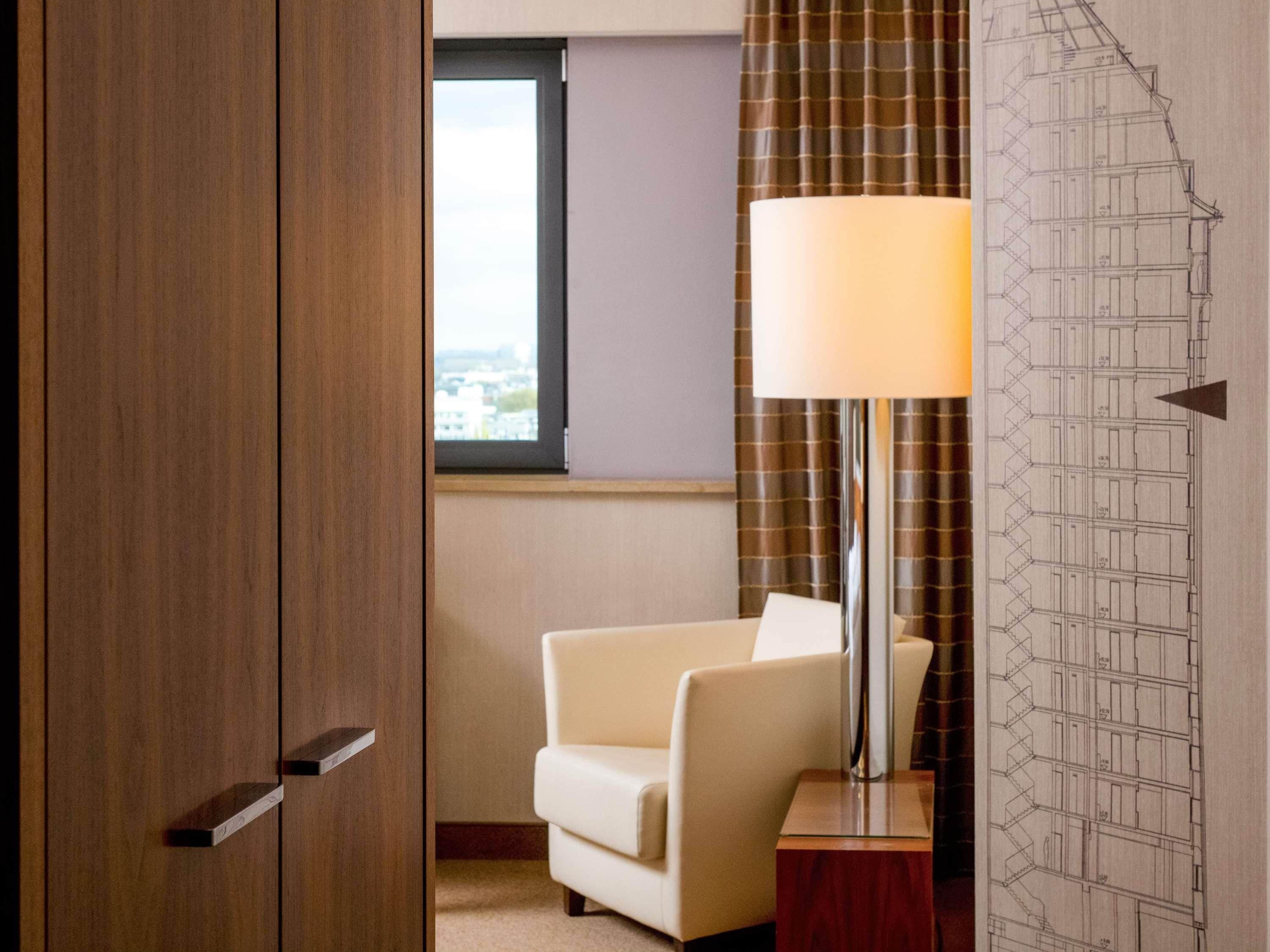 Una habitación de hotel con un armario de madera, un sillón blanco, una lámpara de pie y una ventana con vistas.
